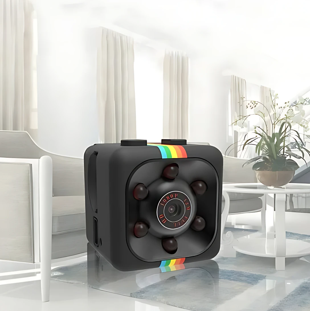 Cámara de seguridad espía en forma de cubo negro con detalles multicolores, lente central y botones laterales, sobre mesa de vidrio en sala moderna.
