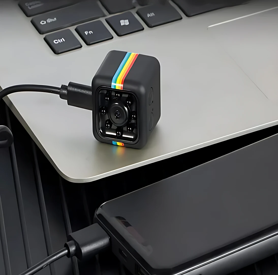 Cámara de seguridad espía negra con franja multicolor, conectada a laptop, ideal para grabar reuniones y clases de forma discreta.