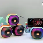 Cuatro altavoces portátiles JK-P60 en colores verde, negro, blanco y azul, con diseño redondo y panel frontal multicolor, acompañados de su caja y un smartphone mostrando video.