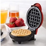 Waflera eléctrica roja abierta con waffle dorado dentro, sobre mesa con fresas y jugo de naranja al fondo.