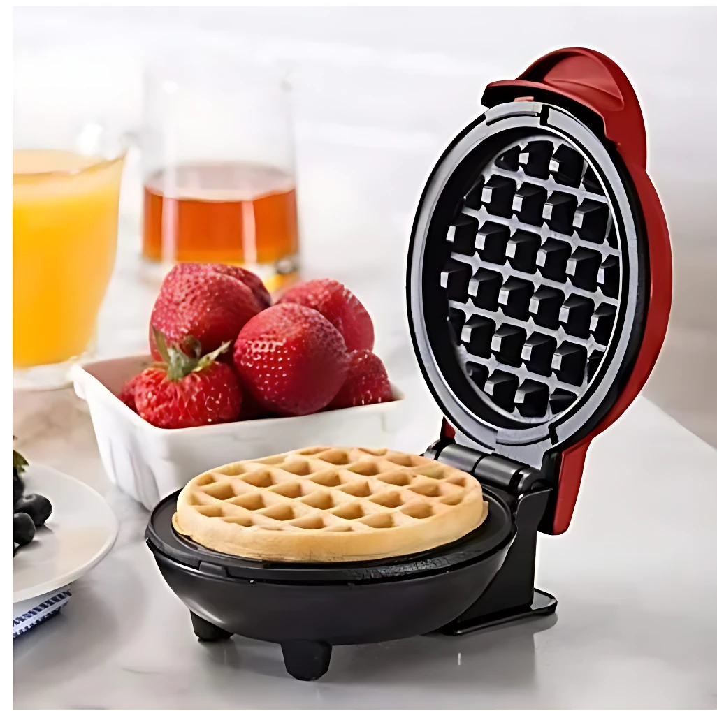 Waflera eléctrica roja abierta con waffle dorado dentro, sobre mesa con fresas y jugo de naranja al fondo.