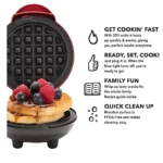 Mini waflera eléctrica roja con waffles dorados y frutos rojos encima, vista frontal mostrando rejilla de cocción y base negra.
