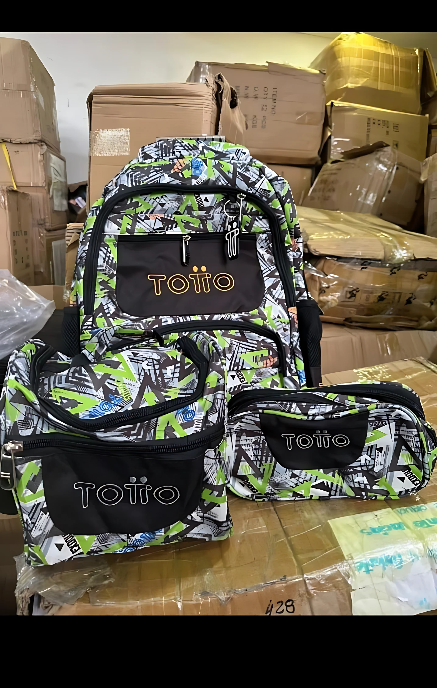 Mochila escolar juvenil tipo Totto con diseño geométrico verde y negro, incluye estuche lateral y bolsa de almuerzo, sobre cajas de cartón en almacén.