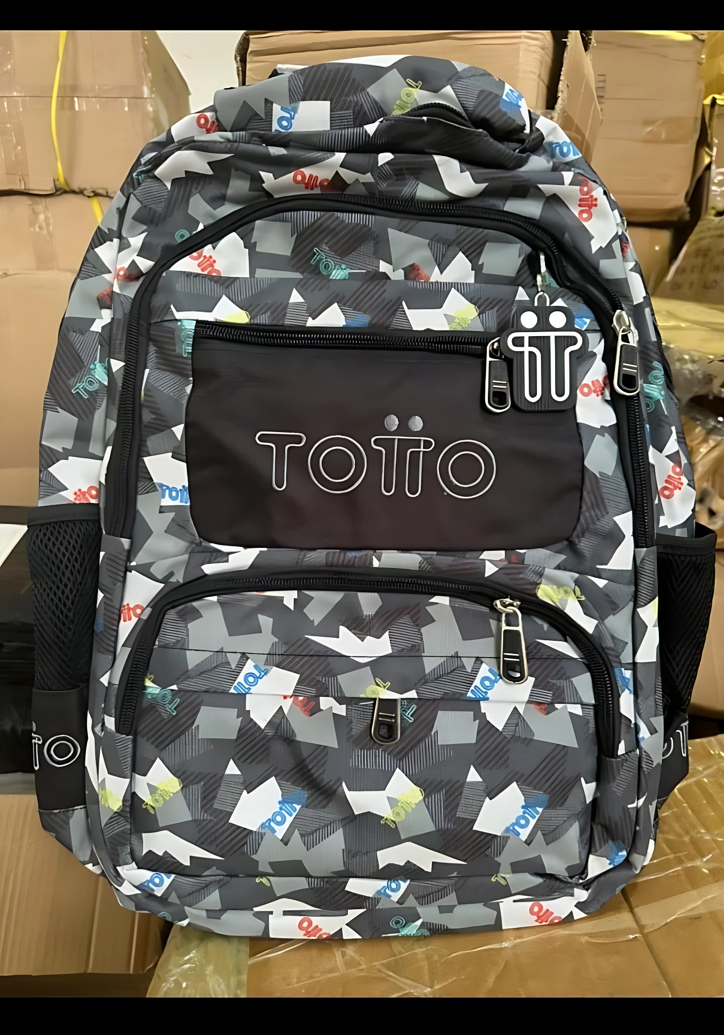 Mochila escolar juvenil tipo Totto con diseño geométrico en gris, blanco y azul; presenta compartimentos frontales con cremalleras y logotipo visible.