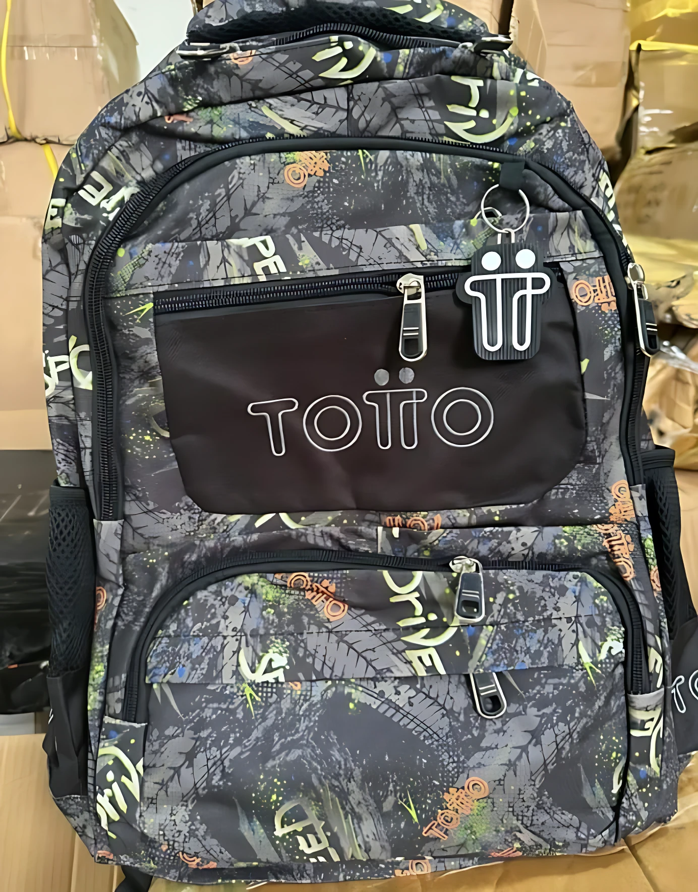 Mochila escolar juvenil tipo Totto con diseño geométrico en tonos oscuros, detalles en verde y naranja, compartimentos frontales con cremalleras y logotipo visible.
