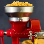 Molino de granos en hierro fundido color rojo con tolva metálica llena de maíz amarillo, mostrando salida de producto procesado.