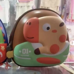 Mochila infantil rígida de color marrón con diseño tridimensional de jirafa en tonos naranjas y verdes, mostrando el frente con detalles gráficos y cierre lateral visible.