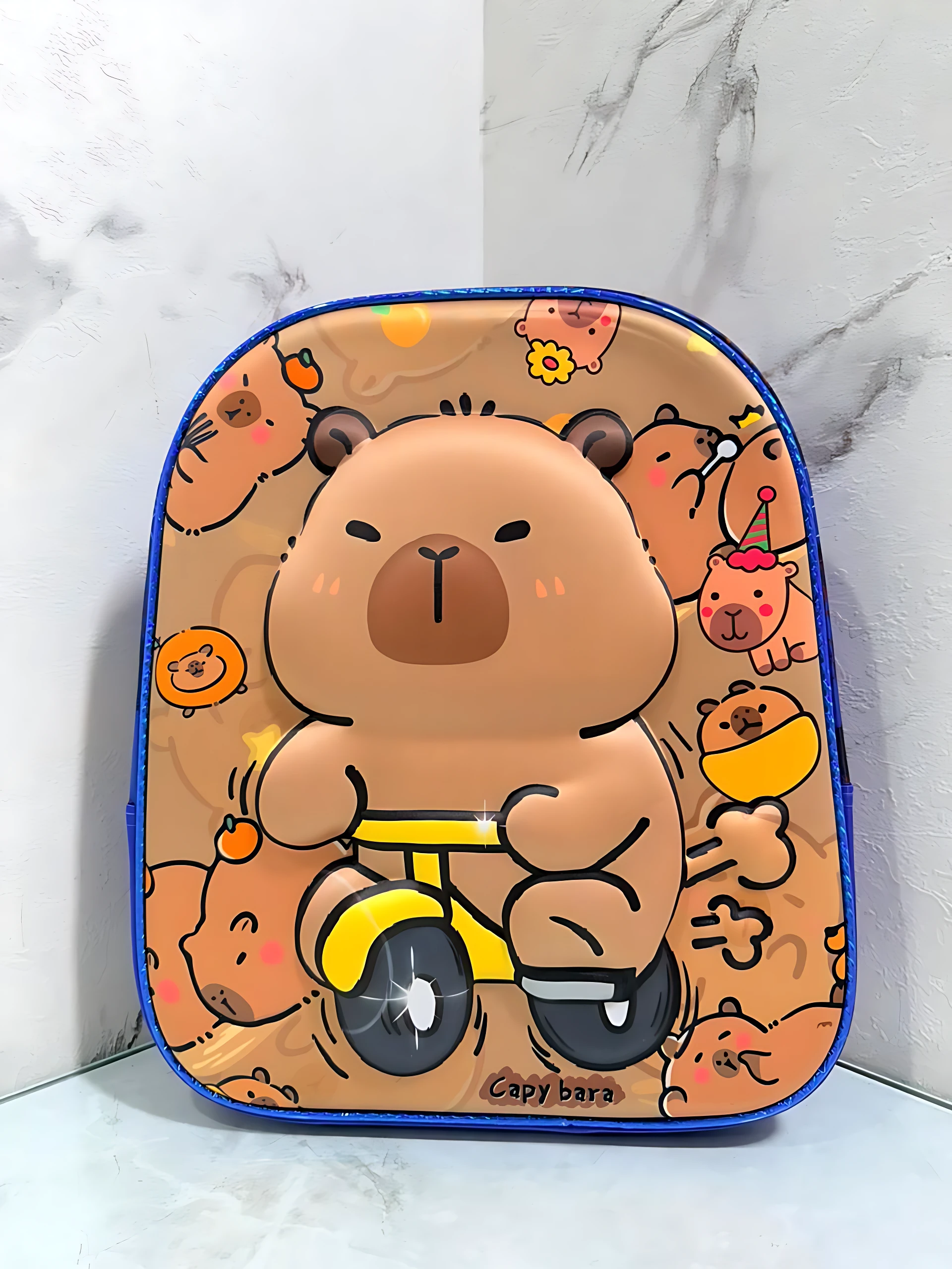 Mochila escolar infantil compacta con cierre único, diseño de capibara montando triciclo amarillo sobre fondo beige con estampado repetido.