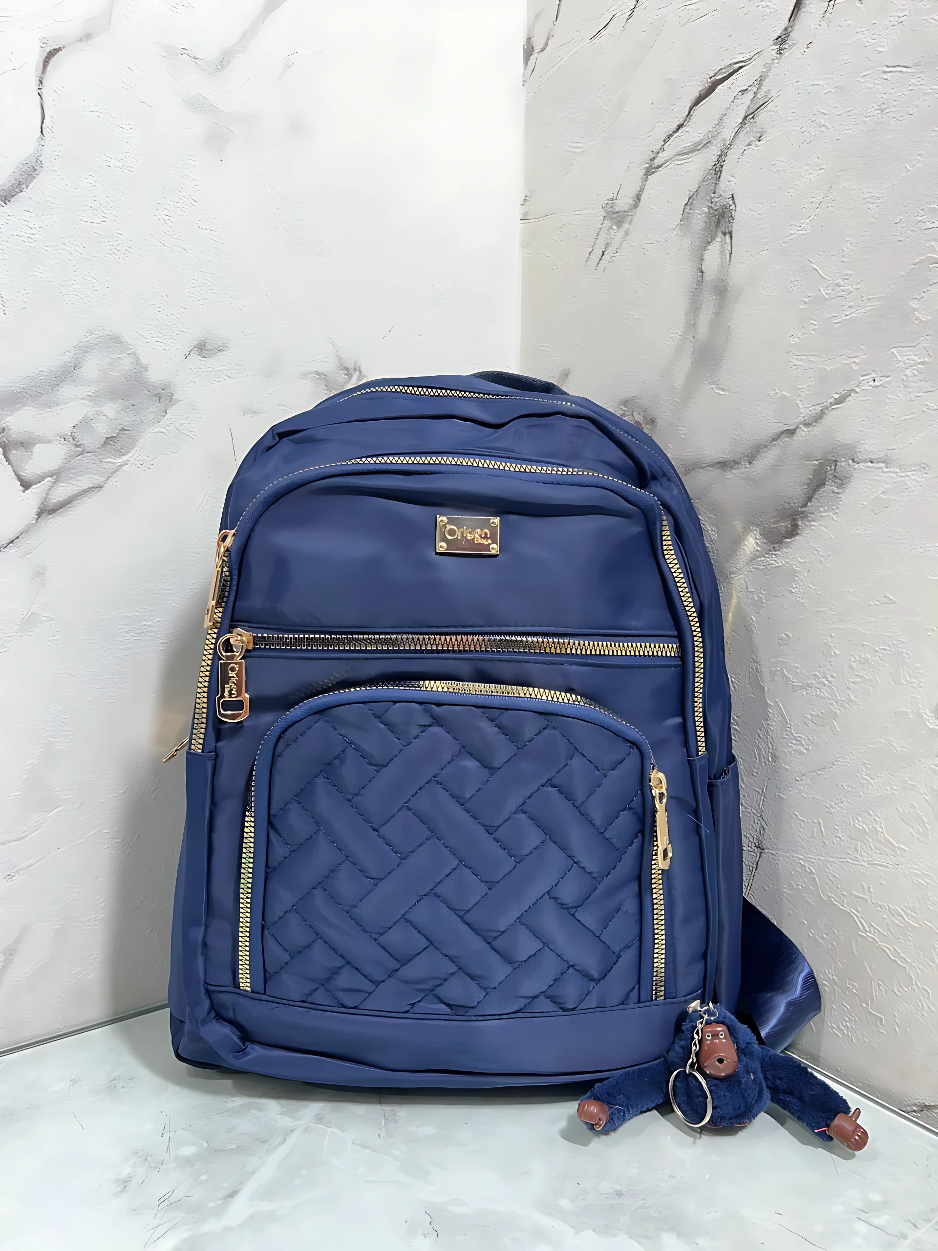 Morral Origen Mn64 en azul marino con costura entrelazada y cremalleras doradas, acompañado de llavero de monedita.