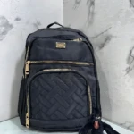 Morral Origen Mn64 en color negro con cremalleras doradas y panel acolchado, acompañado de un pequeño juguete de peluche negro.