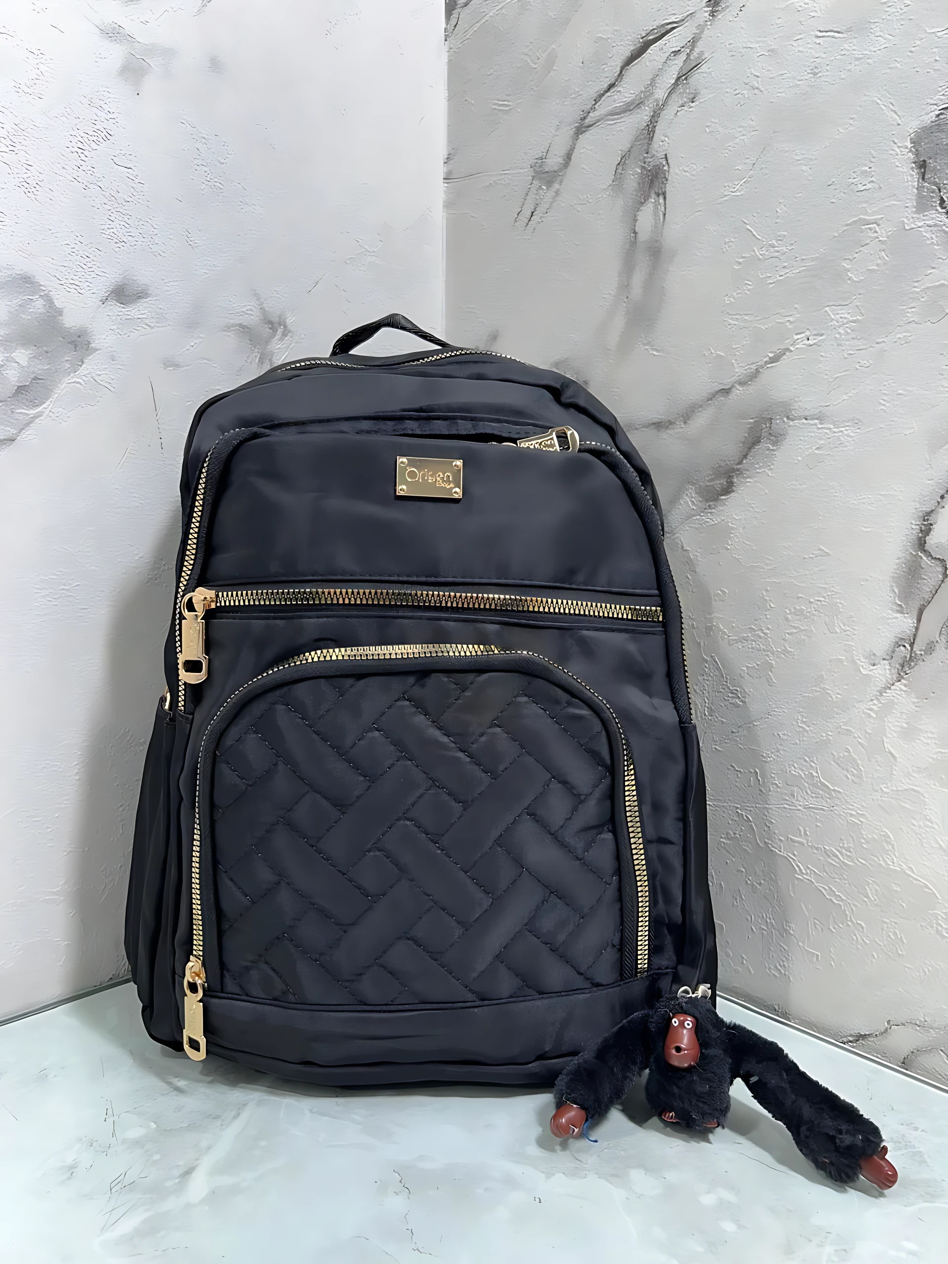 Morral Origen Mn64 en color negro con cremalleras doradas y panel acolchado, acompañado de un pequeño juguete de peluche negro.