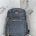 Morral Origen Mn64 en gris oscuro con costuras entrelazadas y cremalleras doradas, mostrando su diseño frontal con múltiples compartimentos y un pequeño peluche gris al pie.