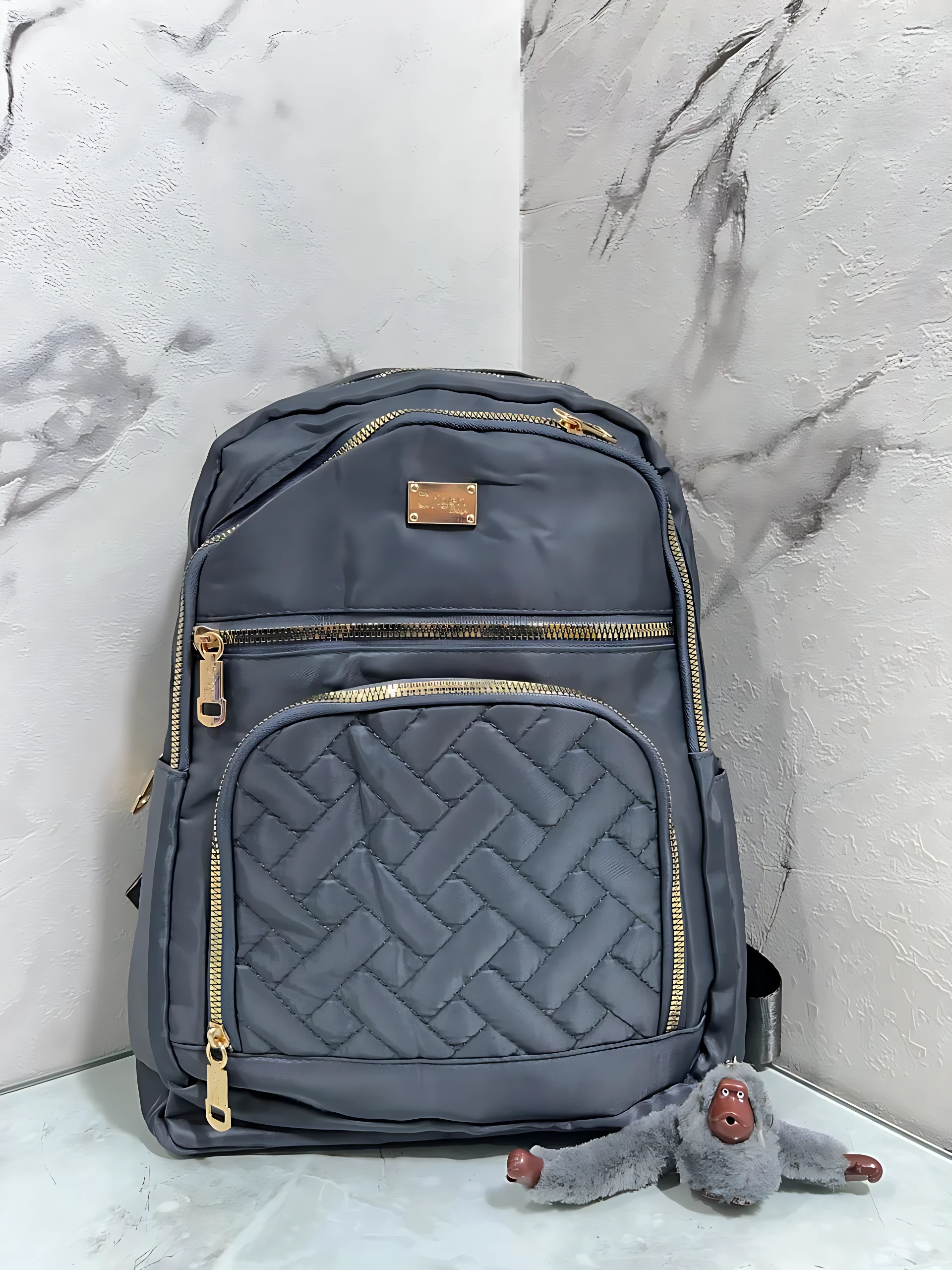 Morral Origen Mn64 en gris oscuro con costuras entrelazadas y cremalleras doradas, mostrando su diseño frontal con múltiples compartimentos y un pequeño peluche gris al pie.