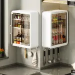 Dos organizadores de pared blancos con puertas transparentes y asas metálicas, montados en esquina de cocina. Muestran estantes internos con frascos y botellas, y ganchos inferiores para colgar utensilios como cuchillos, espátulas y trapo.