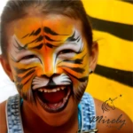 Niña sonriente con maquillaje facial de tigre en tonos naranjas, negros y blancos, mostrando diseño detallado con bigotes y dientes; fondo amarillo con rayas negras.