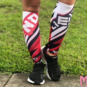 Pantorrilleras de compresión graduada en diseño bicolor rojo y negro con detalles blancos, usadas sobre calzado deportivo, sobre césped.