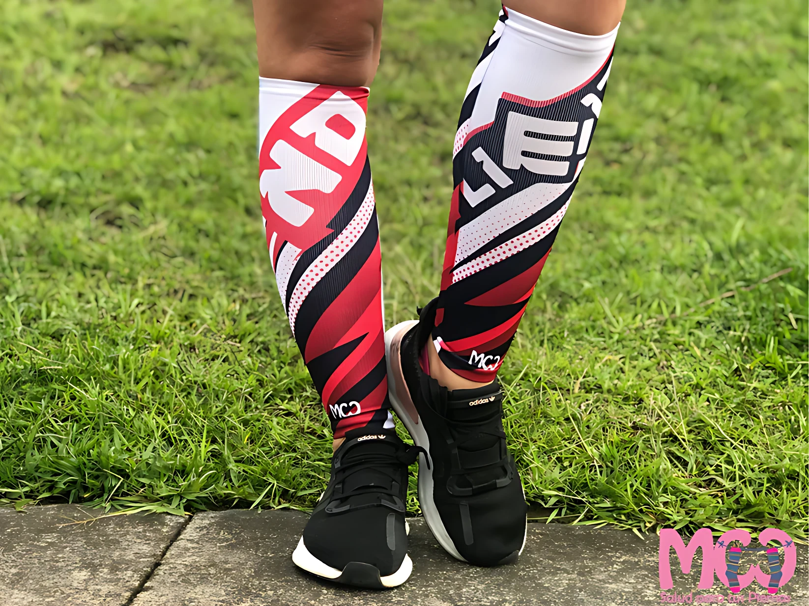 Pantorrilleras de compresión graduada en diseño bicolor rojo y negro con detalles blancos, usadas sobre calzado deportivo, sobre césped.