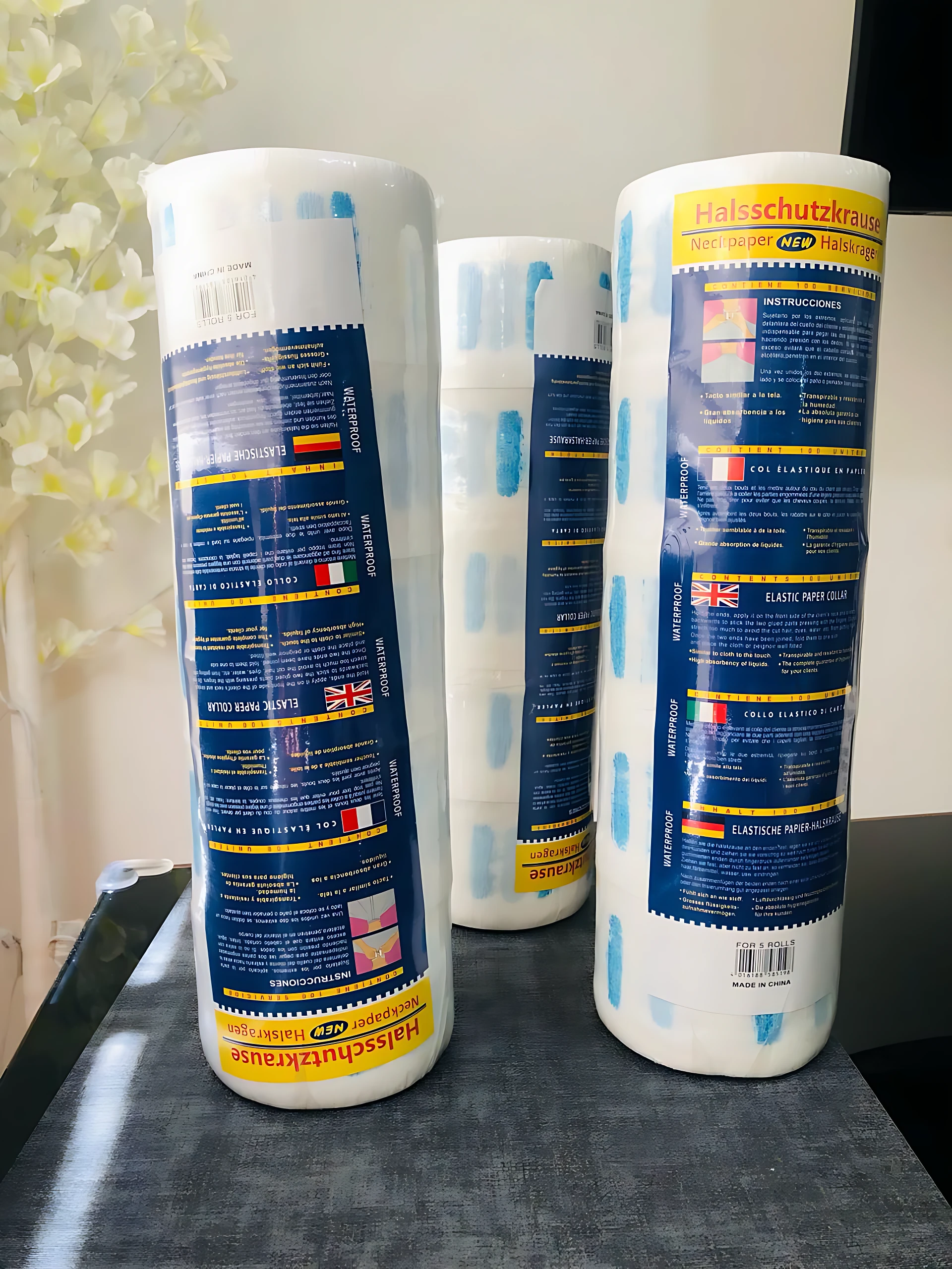Tres rollos de papel cuello para barbería con adhesivo resistente, presentados en empaque blanco y azul, sobre superficie oscura con fondo decorativo floral.