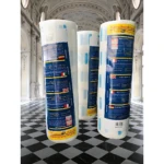 Dos rollos de papel cuello para barbería con adhesivo resistente, presentados en empaque blanco y azul con etiquetas multilingües, sobre piso de baldosas negras y blancas en interior de salón elegante.