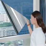 Mujer aplicando papel polarizado plateado en ventana, reduciendo brillo exterior y protegiendo interiores de calor y luz solar.