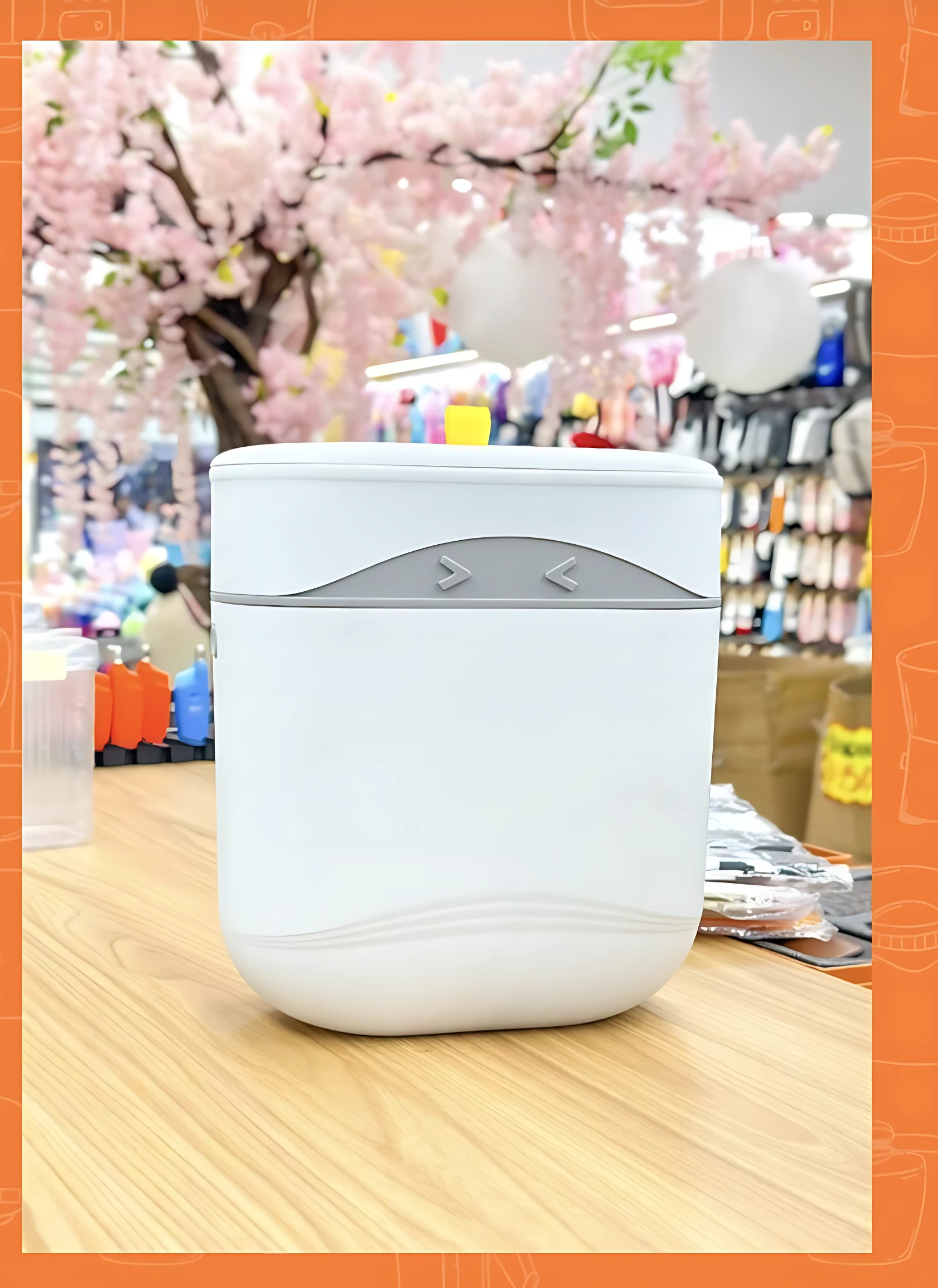 Papelera de cocina blanca con tapa y detalles grises, vista frontal sobre superficie de madera, en entorno de tienda con decoración floral.