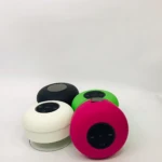 Cuatro altavoces Bluetooth redondos para ducha en colores negro, blanco, verde y rosa, con superficie mate y panel de control visible en la parte superior.
