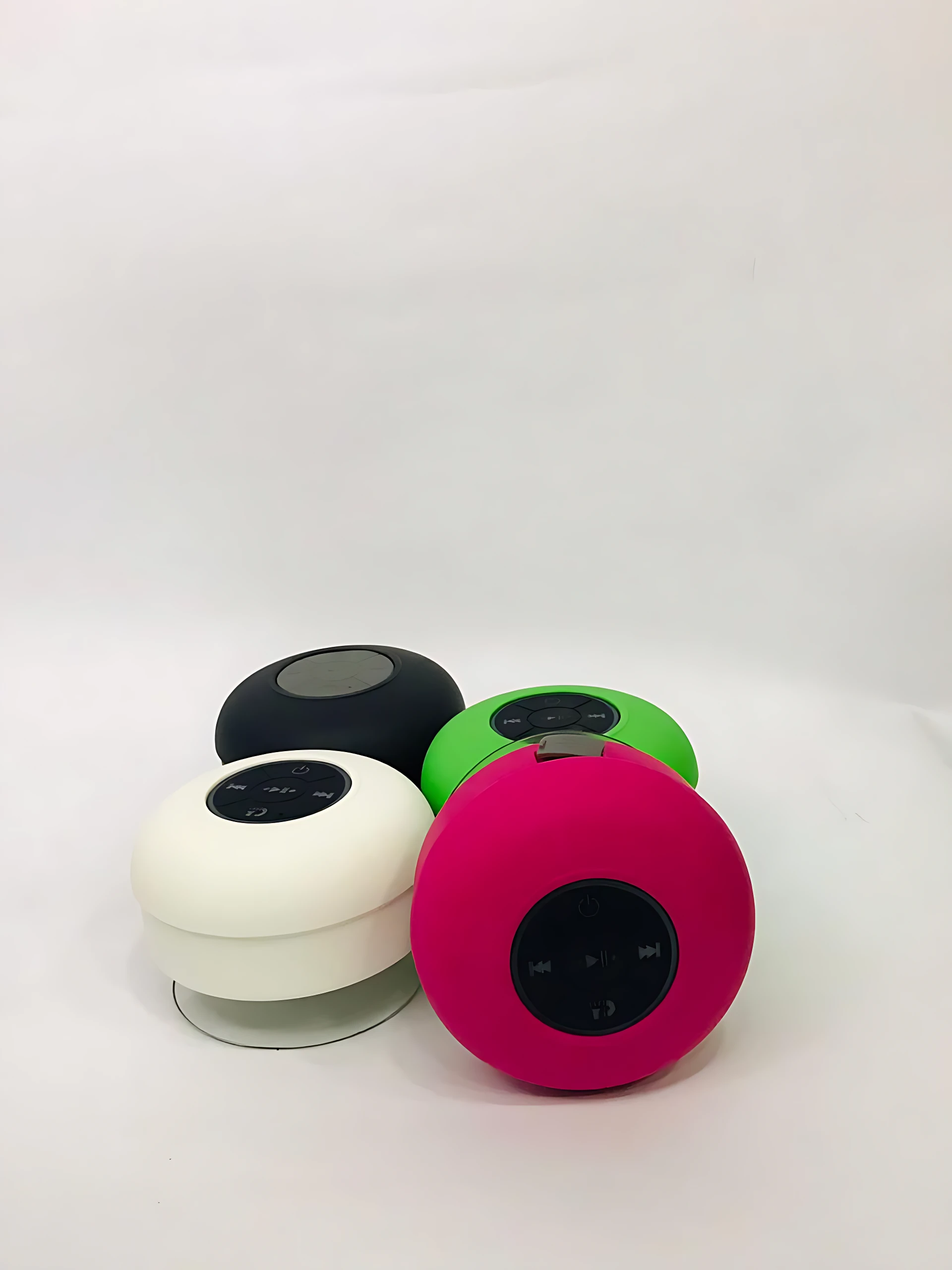Cuatro altavoces Bluetooth redondos para ducha en colores negro, blanco, verde y rosa, con superficie mate y panel de control visible en la parte superior.