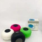 Cuatro altavoces Bluetooth redondos en blanco, rosa, verde y negro, con controles táctiles visibles, junto a su caja de presentación.