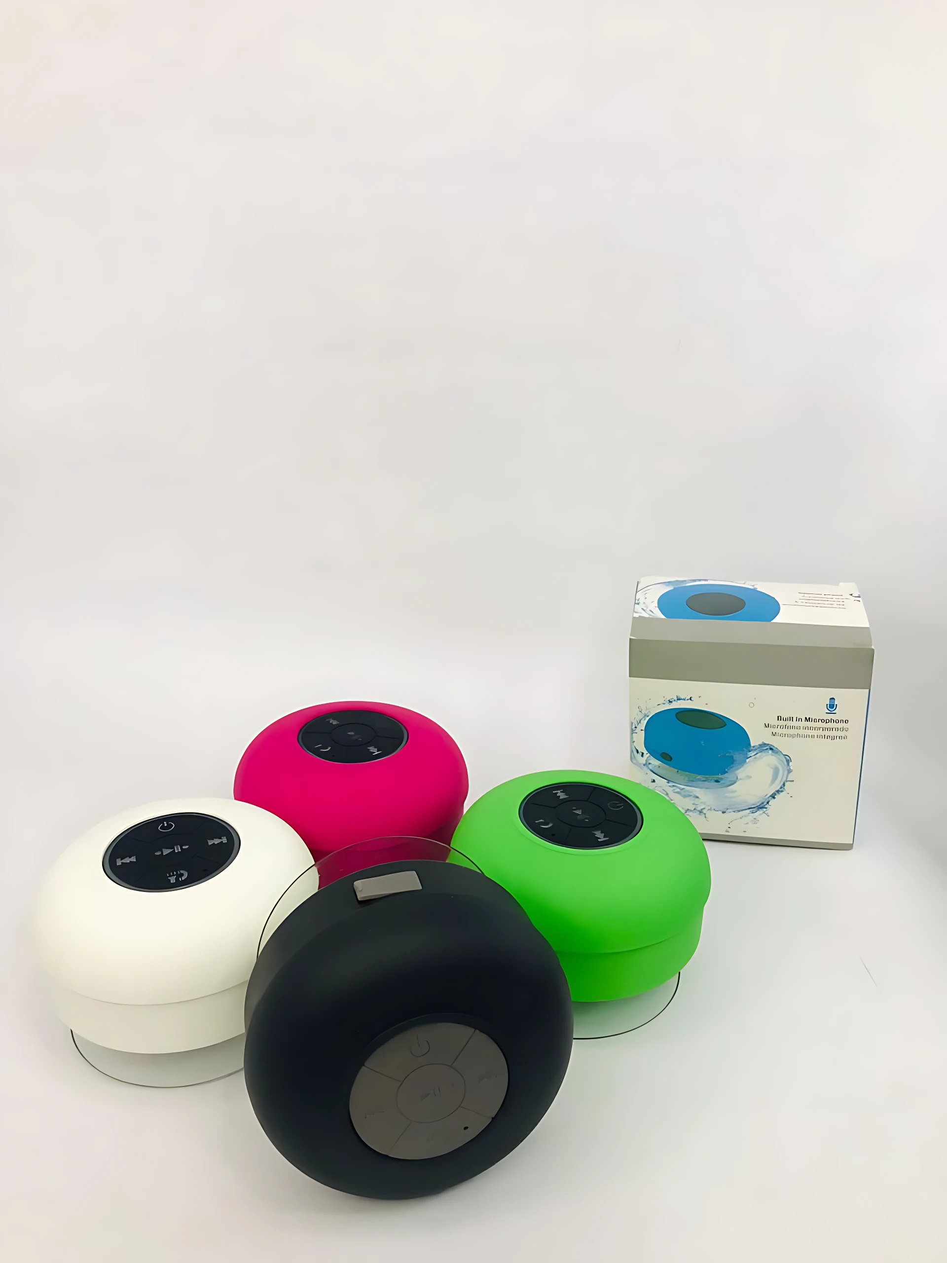 Cuatro altavoces Bluetooth redondos en blanco, rosa, verde y negro, con controles táctiles visibles, junto a su caja de presentación.