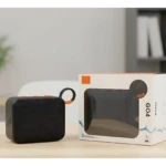 Parlante Bluetooth G04 negro compacto con asa naranja, junto a su caja de venta al por menor sobre superficie de madera.