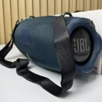 Altavoz JBL Xtreme 4 en color azul oscuro, con textura de malla y correa negra para transporte, mostrando el altavoz frontal y lateral sobre una superficie blanca.