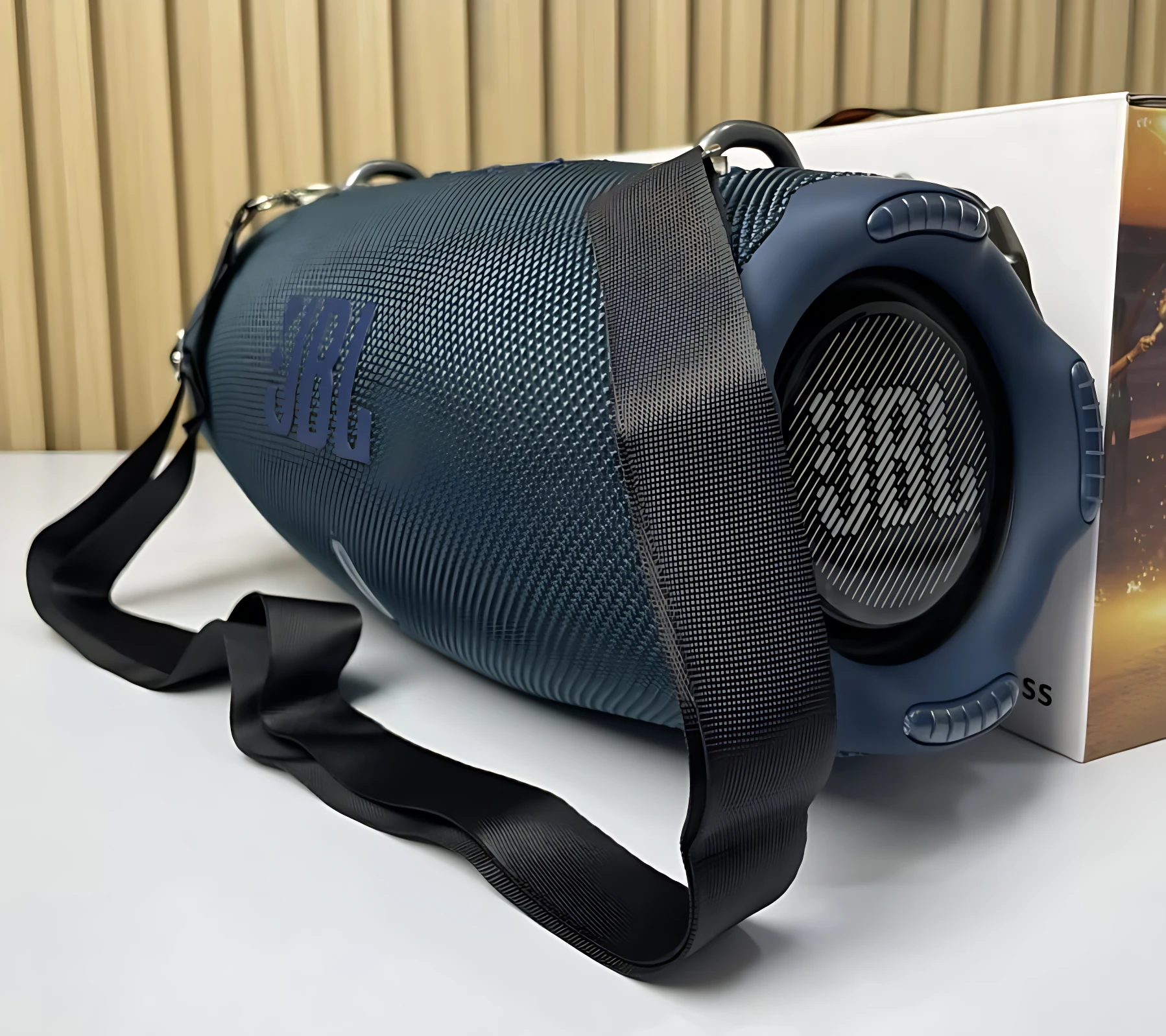 Altavoz JBL Xtreme 4 en color azul oscuro, con textura de malla y correa negra para transporte, mostrando el altavoz frontal y lateral sobre una superficie blanca.
