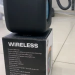 Parlante Bluetooth negro con asa trenzada y detalles naranjas, sobre su caja que indica especificaciones técnicas y colores disponibles.
