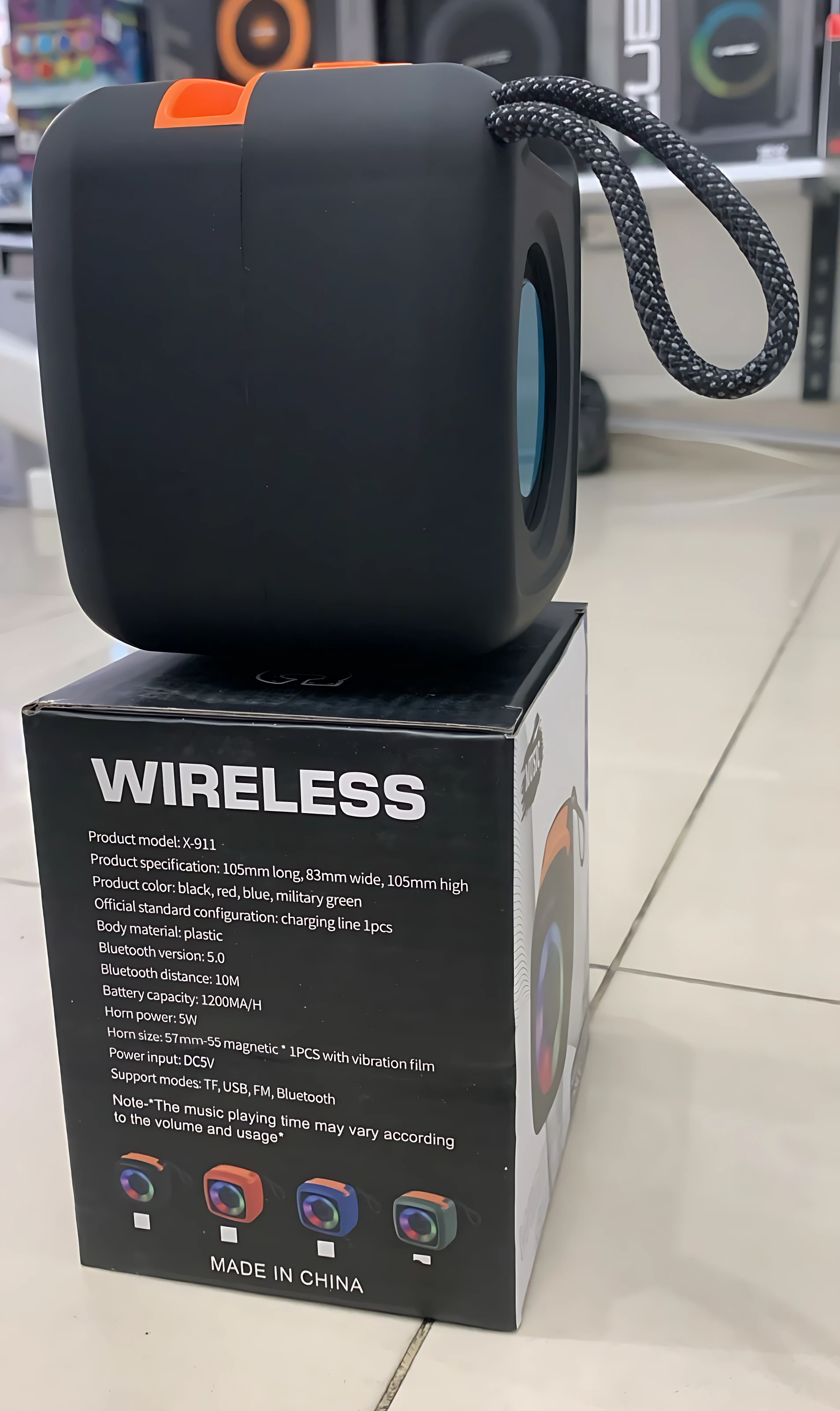 Parlante Bluetooth negro con asa trenzada y detalles naranjas, sobre su caja que indica especificaciones técnicas y colores disponibles.
