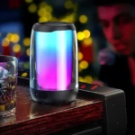 Parlante Remax Bluetooth con luces LED multicolor en ambiente nocturno, sobre superficie de madera junto a vaso de bebida.