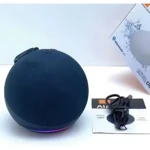 Parlante inteligente esférico negro con luz LED púrpura en base, cable USB y caja visible al fondo.