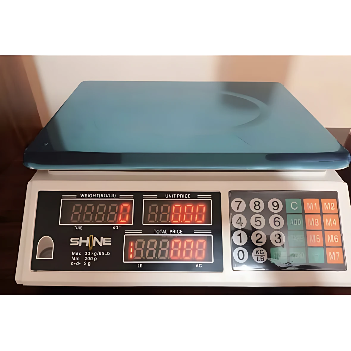 Báscula electrónica de mesa blanca con plataforma azul y teclado numérico. Pantalla LED roja muestra peso, precio unitario y total. Modelo SHINE, capacidad 40 kg.