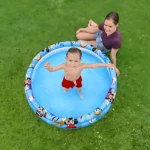 Vista cenital de niño en piscina inflable azul con diseño de Mickey Mouse, sobre césped verde, con mujer sentada al lado.