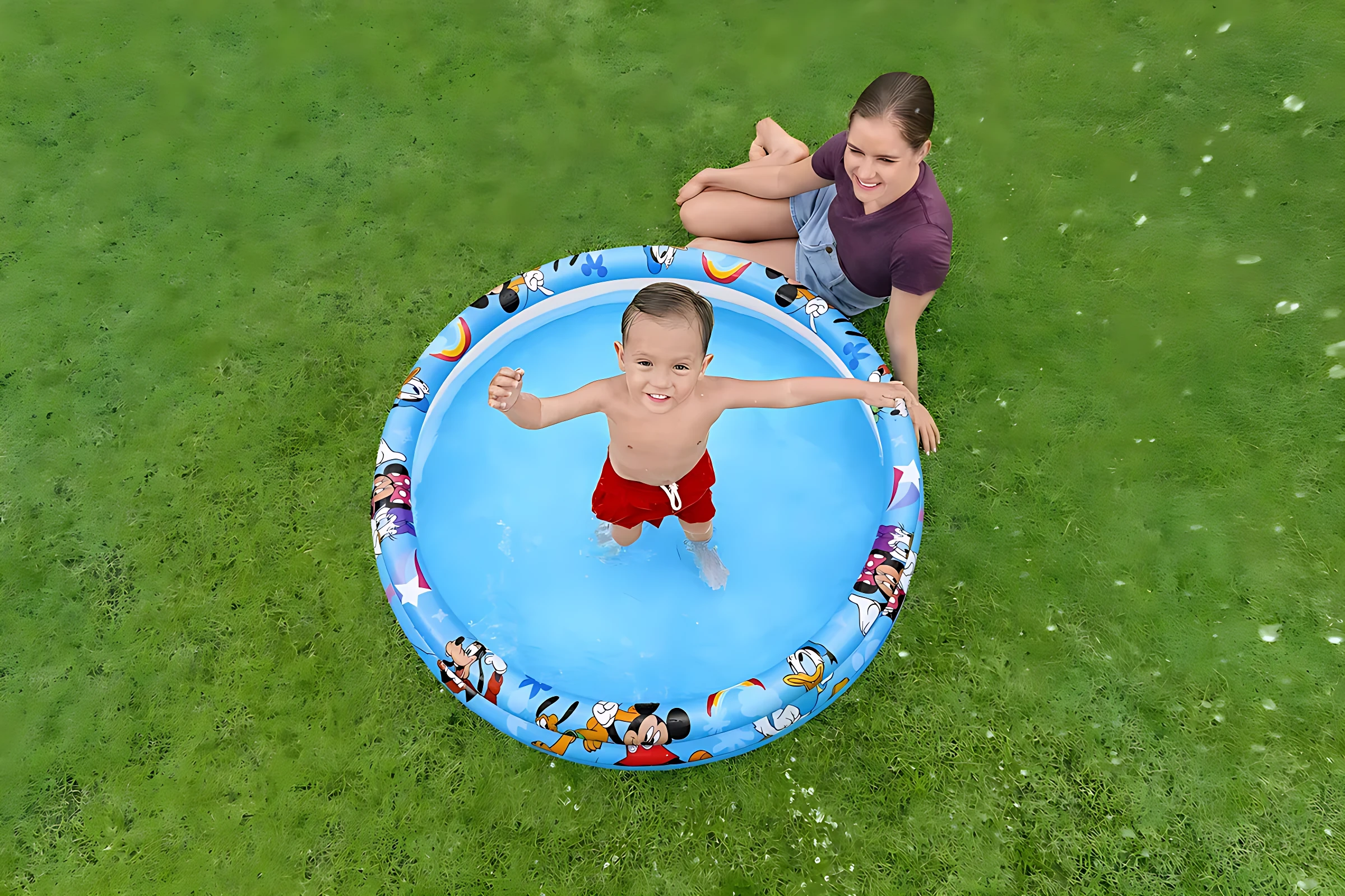 Vista cenital de niño en piscina inflable azul con diseño de Mickey Mouse, sobre césped verde, con mujer sentada al lado.