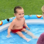 Niño sonriente en piscina inflable azul con anillos de Mickey Mouse, sobre césped verde.