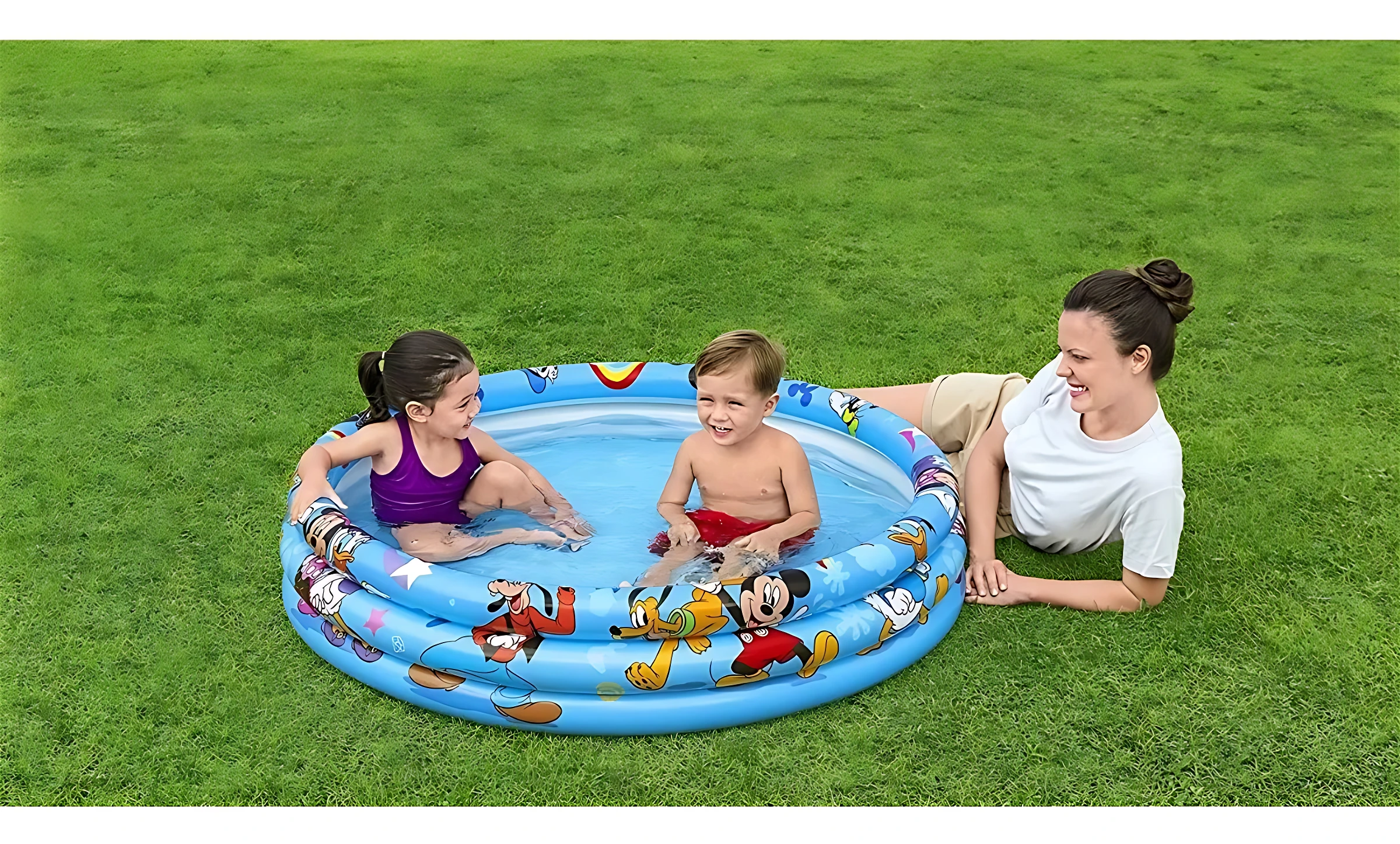 Piscina inflable azul con Mickey Mouse, tres anillos y dibujos de personajes. Niño y niña jugando dentro, mujer acostada al lado en césped verde.