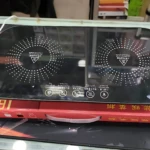 Plato eléctrico calentador de dos zonas, superficie negra con indicadores de temperatura y controles táctiles, sobre caja roja con precio visible.
