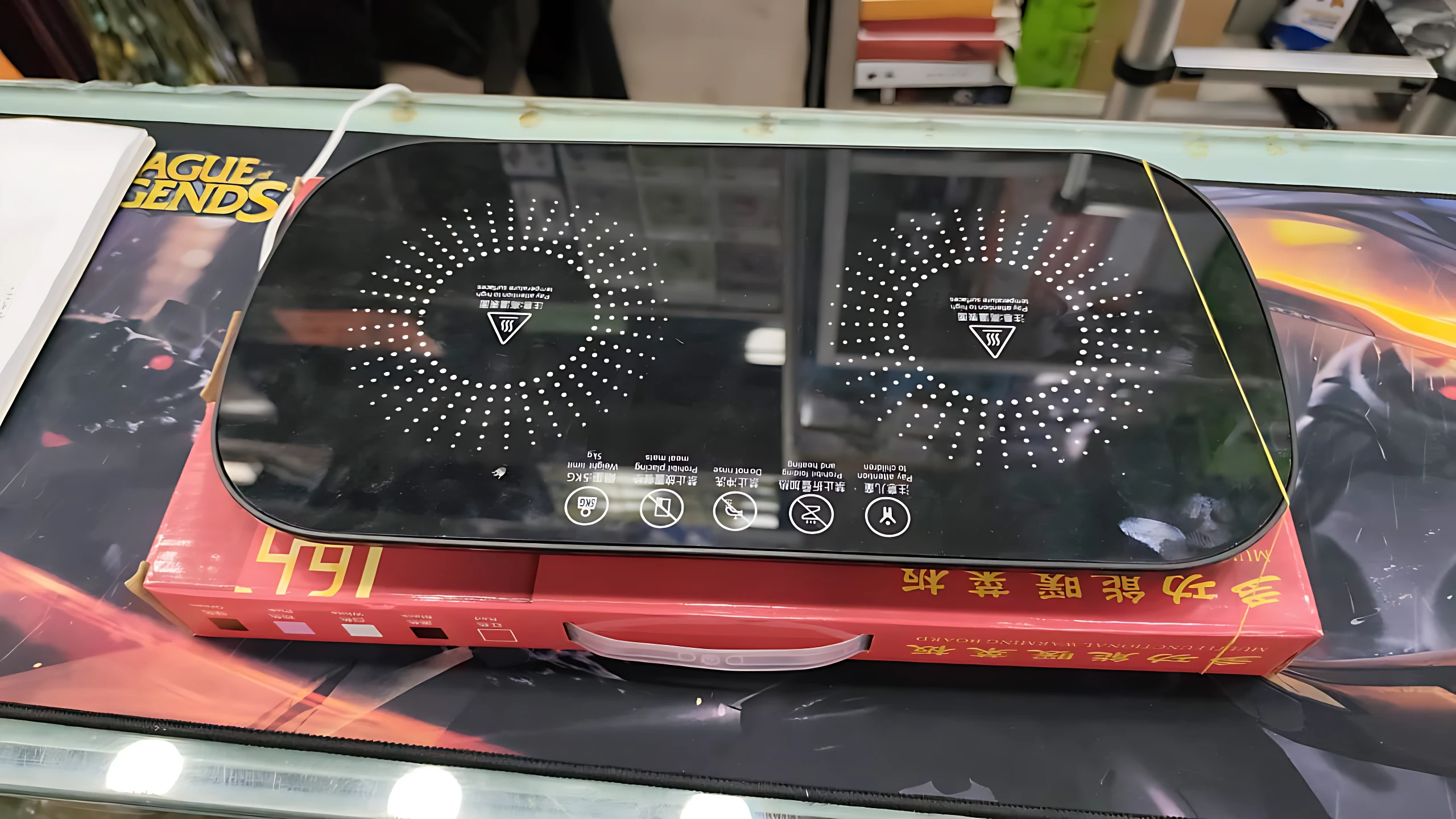 Plato eléctrico calentador de dos zonas, superficie negra con indicadores de temperatura y controles táctiles, sobre caja roja con precio visible.