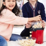 Niña y niño sacan palomitas de cine de una crispetera eléctrica roja sin aceite, con palomitas cayendo a un bowl transparente sobre alfombra blanca.