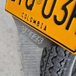 Porta placa de acero inoxidable para Suzuki GN 125, instalado en la parte trasera de la moto con matrícula amarilla colombiana visible.