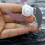 Procesador de alimentos portátil inalámbrico con cuchillas metálicas y base blanca, mostrando detalle de la pieza de corte en mano sobre superficie oscura.