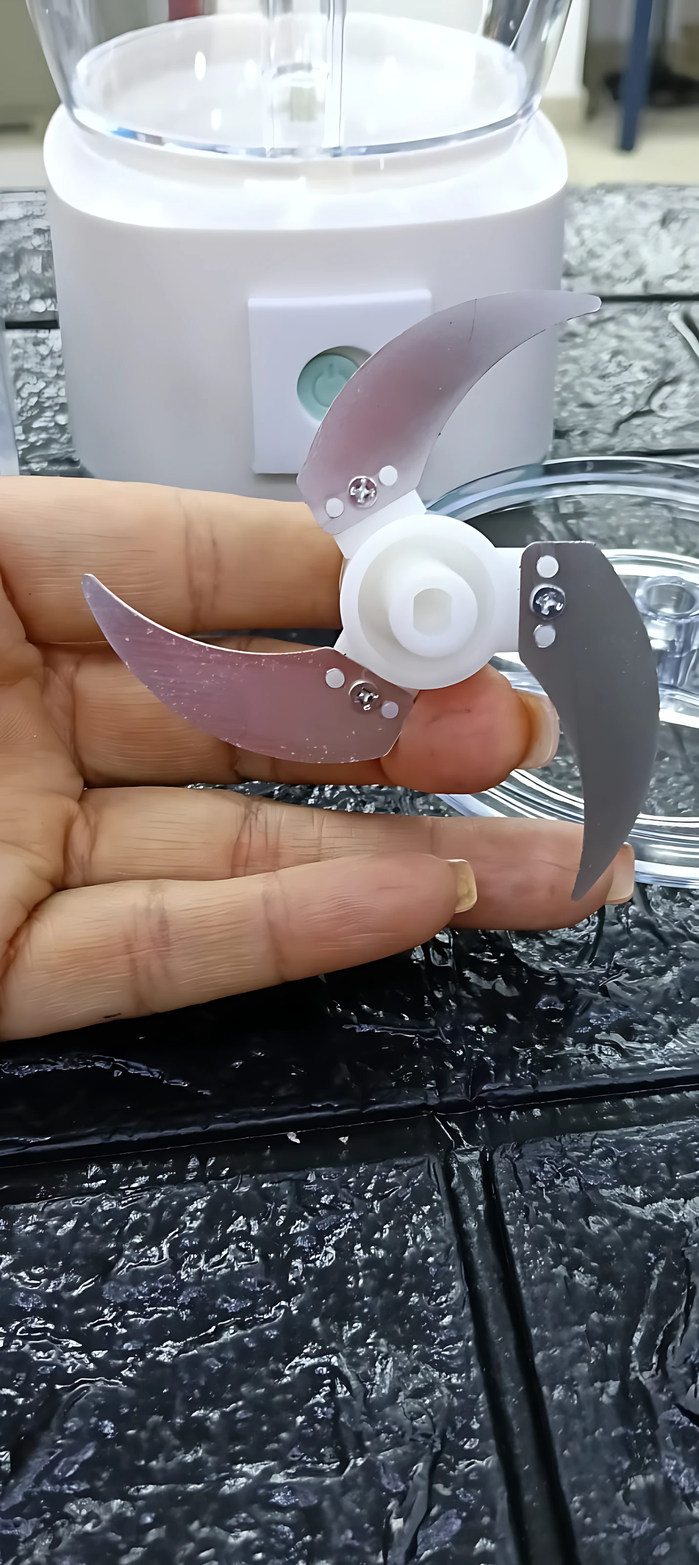 Procesador de alimentos portátil inalámbrico con cuchillas metálicas y base blanca, mostrando detalle de la pieza de corte en mano sobre superficie oscura.