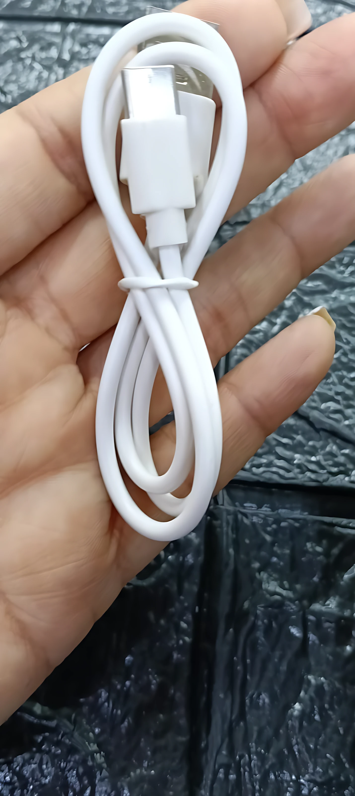 Cable de carga blanco enrollado en mano, con conector USB tipo A y micro USB, sobre fondo texturizado oscuro.