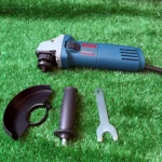 Pulidora angular Bosch GWS 6-115 en color azul sobre césped artificial, acompañada de su protector, soporte y llave de ajuste.