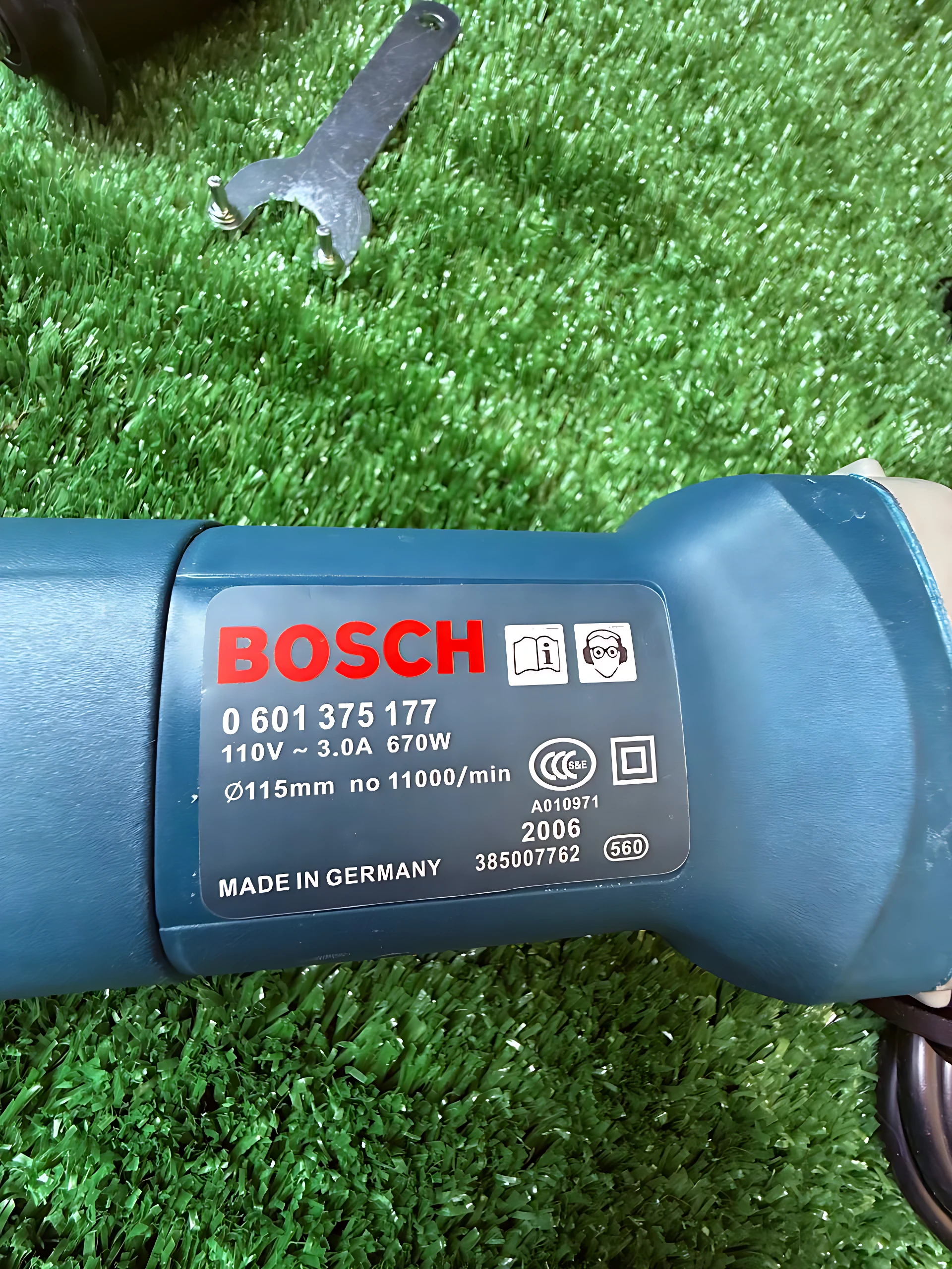 Etiqueta de Bosch en pulidora azul sobre césped artificial, con datos técnicos visibles: 670W, 11000 rpm, Ø115mm, modelo 0 601 375 177, fabricada en Alemania.